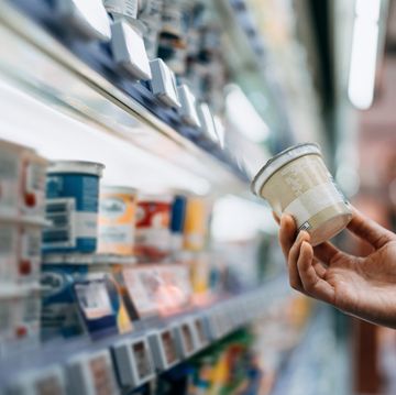 close up of a woman's hand shopping for fresh organic healthy yoghurt along the dairy aisle in supermarket, reading the nutrition label on the pot. routine grocery shopping. healthy eating lifestyle. making healthier food choices. smart eating concept