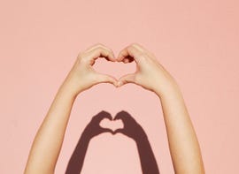 a hand making a heart shape in front of a pink wall with a shadow of the hand