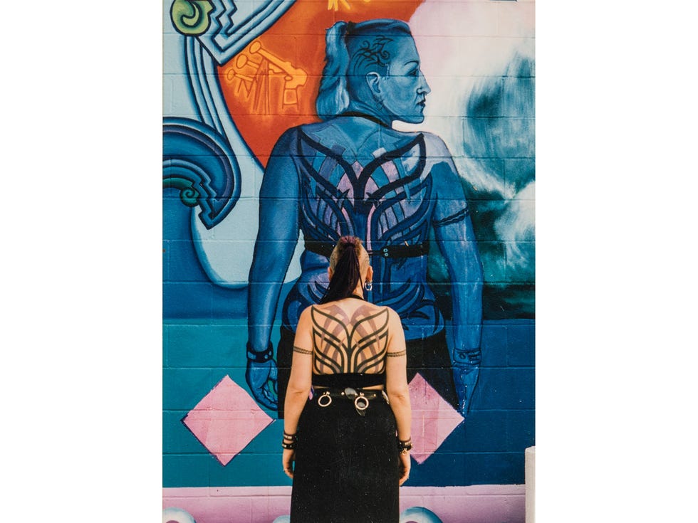ATA080918murals Img02 Model posing in front of detail from "La Historia de Adentro/La Historia de Afuera" by Yreina D. Cervantez and Alma Lopez, 1995. The mural focuses on the contributions of the minority communities of Huntington Beach to its history. Painted on the parking lot wall of the Huntington Beach Art Center, it drew from the life stories of residents, some of whom served as models. The artists’ distinct perspective on local history was erased in 2009, when the mural was whitewashed by a new building owner.