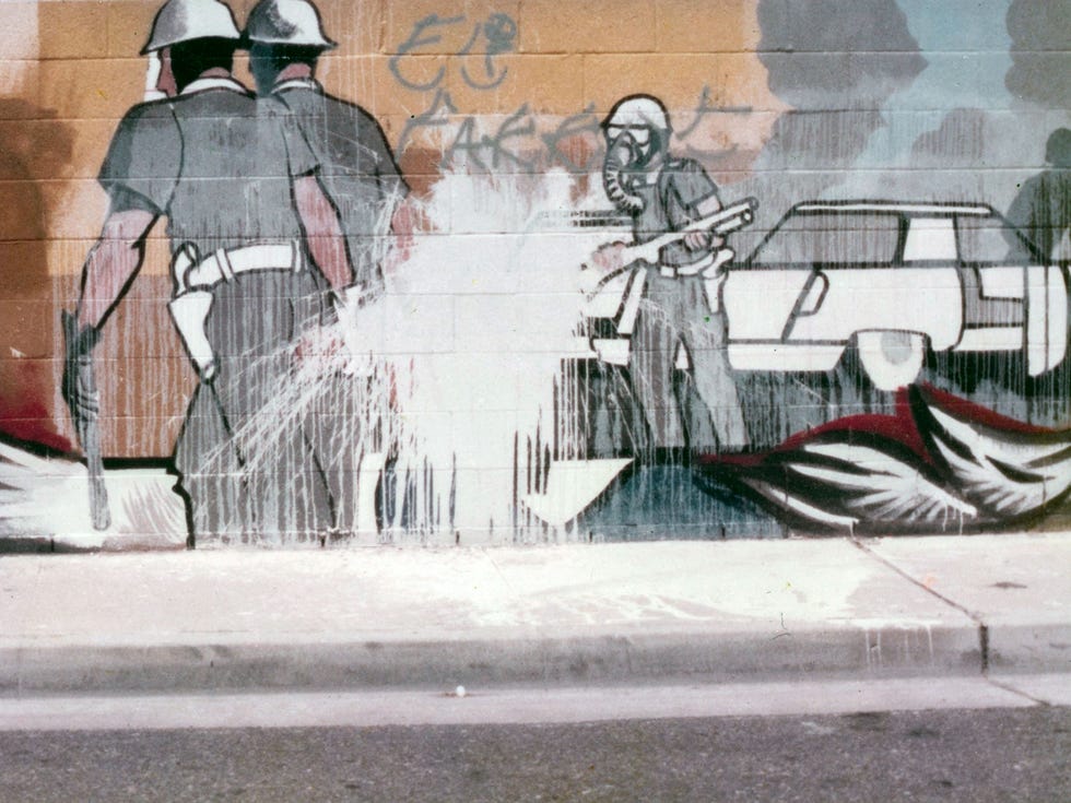 ATA080918murals Img03 Detail from the "Fountain Valley Mural." Early in the mural-making process, a controversy arose with the Fountain Valley Police Department over a scene depicting a young man’s arrest by police dressed in riot gear. The controversy received a significant amount of media attention, and during the dispute, someone threw a bucket of white paint at the scene. O’Cadiz chose to keep the white paint on the mural as a permanent mark of opposition and evidence of the controversy, which ultimately lead to loss of funding for the project.