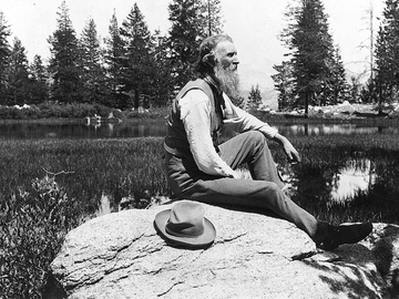 John Muir seated on rock with lake and trees in background.