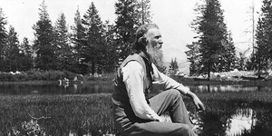 John Muir seated on rock with lake and trees in background.