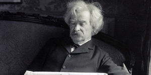 Portrait photograph of Samuel Langhorne Clemens, more popularly known as Mark Twain, seated, reading a newspaper.