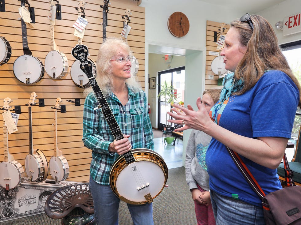 In the display room at the Deering Banjo Company Janet Deering speaks to visitors Jennifer Allison and daughter Aislyn, 8. They and other family members there are from Tucson, Arizona and are about to take a tour of the facility. Janet is holding an Ivanhoe Gold model. At left are the company's Goodtime line of banjos.