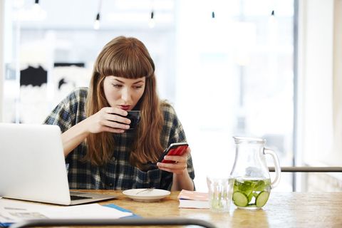 Ragazza alla scrivania davanti a un portatile che beve un caffè e guarda sullo smartphone le offerte di lavoro