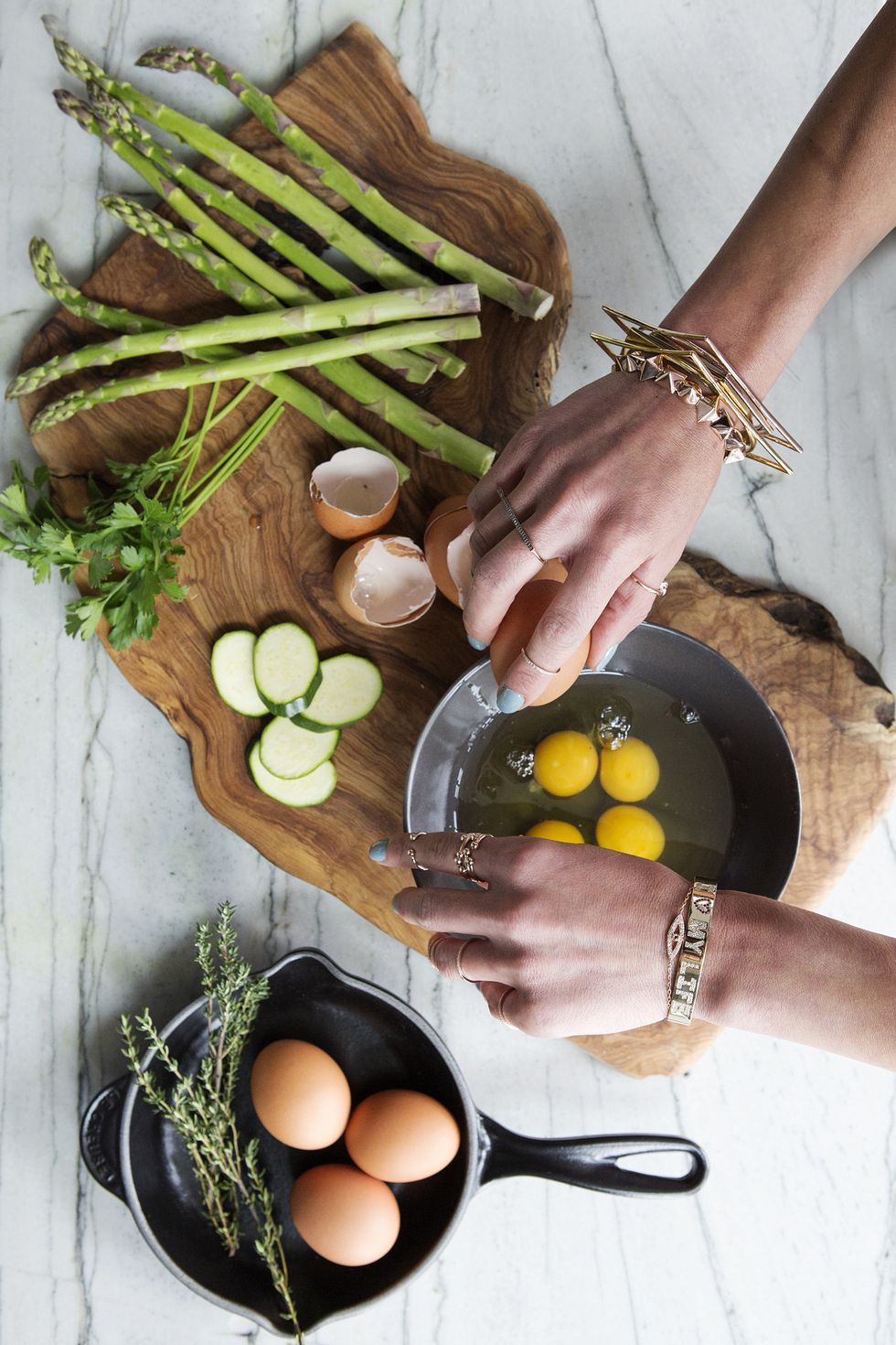 A young woman's hands with jewelry on is breaking eggs into a brown bowl. There are various ingredients for an omelet or frittata. An overhead shot on a marble background.
