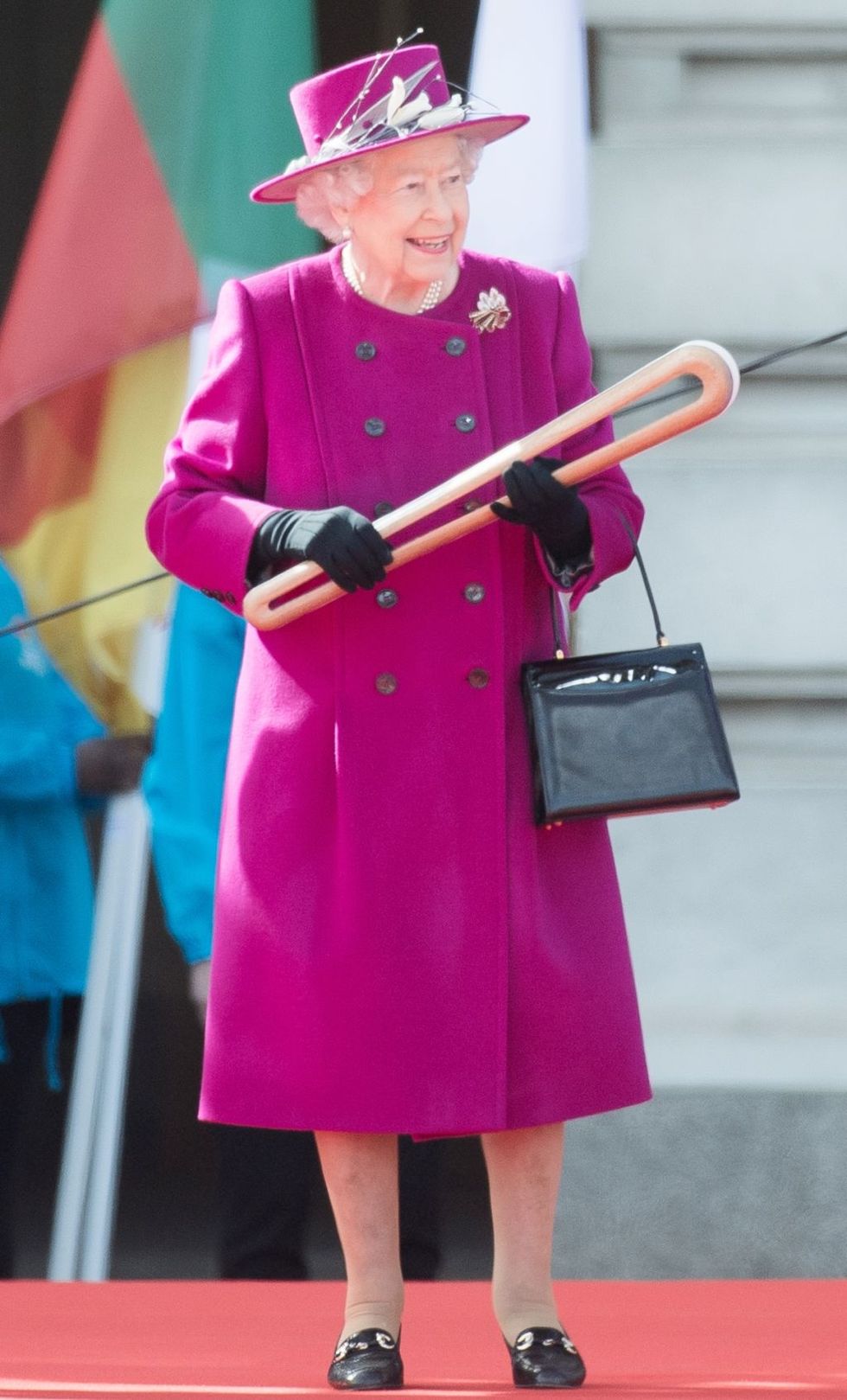<p>印象的なマゼンタピンクは、エリザベス女王にとってシグネチャー的存在であるカラーのひとつ。ポイントはハットの装飾が寒色のシルバーグレーという点。黒い小物と相まって全体がスッキリとした印象に。手に持っているのは、イギリスで4年に1度あるオリンピックのようなスポーツ大会「コモンウェルスゲーム」の聖火リレー、クイーンズバトンリレーのバトン。バトンの中には女王から選手へのメッセージが入っているそう！</p>