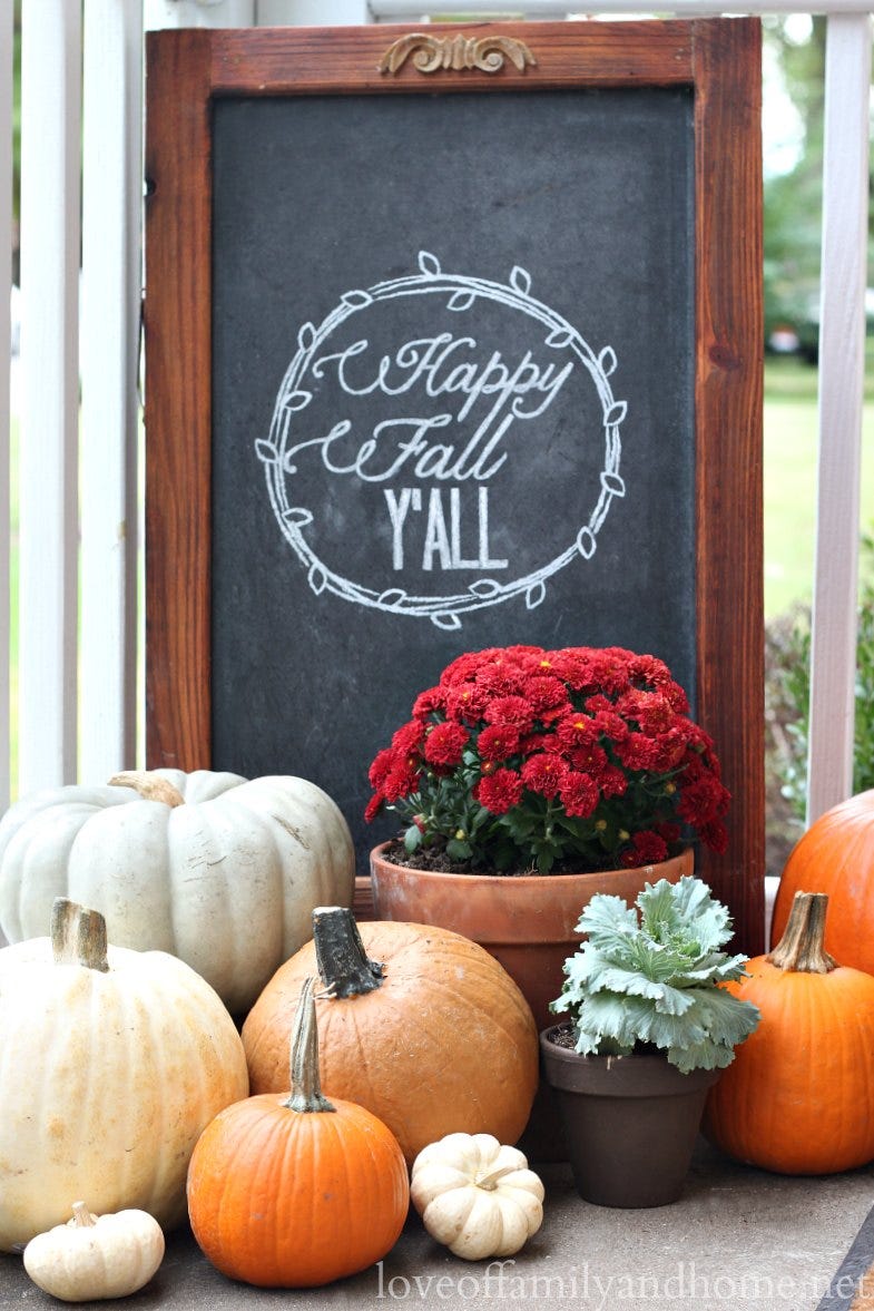 front porch autumn decor with red mums, potted kale, pumpkins, and wood framed chalkboard sign reading happy fall y'all