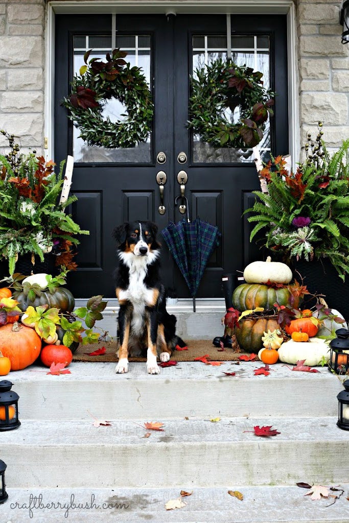 stone house with porch and black double front doors decorated for fall with stacks of pumpkins, wreaths, lanterns