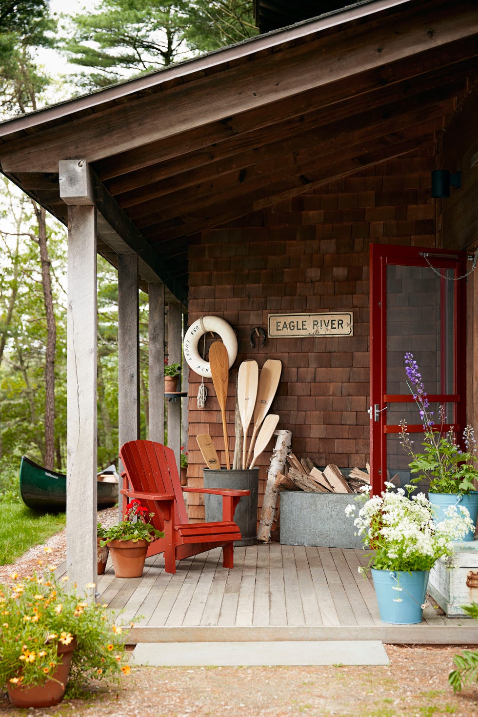 lake cabin front porch with red adirondack chair and plain wooden paddles corralled in metal container along with firewood