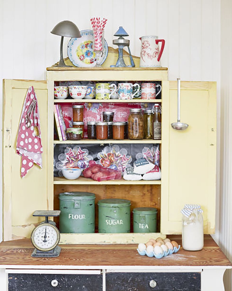 an old yellow shelf rests on top of a worktable and is filled with baking supplies and coffee mugs and jars of jams