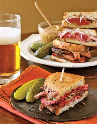 pastrami sandwich halves and pickles on plates next to a glass of beer