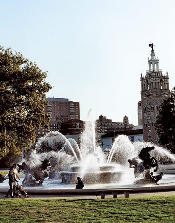 large fountain with iron sculptures in front of a tall tower and buildings