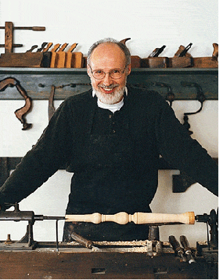 man smiling in front of a wood lathe with sculpted chair leg