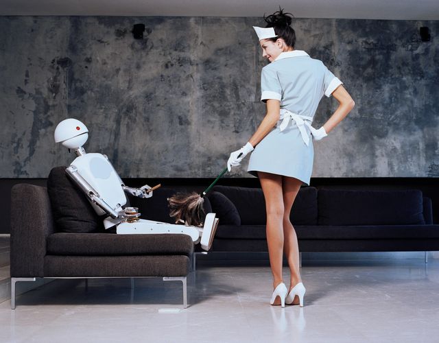 A woman dusting while a robot sits in a chair and drinks whisky