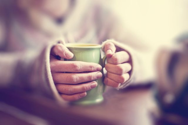 Green mug of hot drink being held in pair of hands of jumpered person