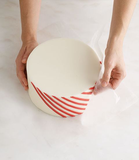 <p>In a bowl, combine 1 Tbsp each light corn syrup and water. Using a paintbrush, brush the stripes with the syrup mixture. Cut the tape off the wax paper. Line up the bottom of the wax paper with the bottom of the cake. Holding the sides of the wax paper, lift the paper, pressing the stripes against and around the cake, making sure that the bottom of the paper is flush with the counter and bottom of the cake. Lightly press on the stripes to make sure they are completely attached. Starting at one corner, carefully peel the wax paper away from the cake (if the stripes start to lift, brush them with the syrup mixture and press onto the cake). Repeat with the template, wax paper and red stripes until the entire cake is covered in stripes. Use a razor blade to gently adjust and straighten stripes immediately after they're placed on the cake.</p>