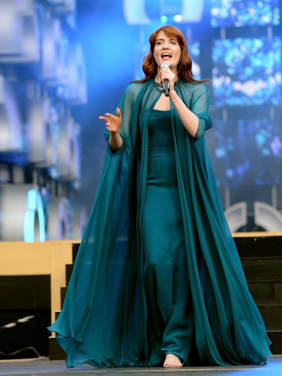 <p>Florence Welch in a deep turquoise silk crepe strapless evening gown with a matching silk chiffon cape featuring a crystal broach closure at the neck.</p>