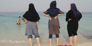 Three women with their trousers rolled up chill out on the shore of Kish Island enjoying lesser restrictions imposed on visitors. Iranian women are not allowed to swim in a bathing suit, their body must be covered at all times while in public | ELLE UK