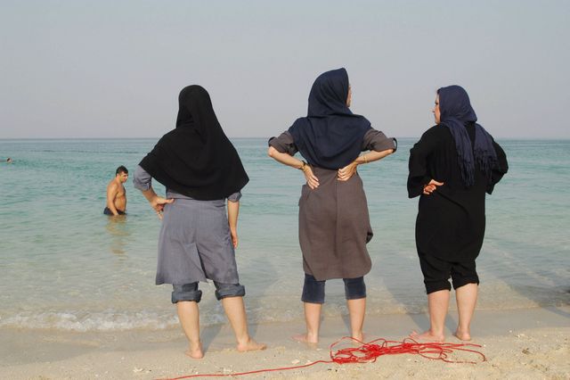 Three women with their trousers rolled up chill out on the shore of Kish Island enjoying lesser restrictions imposed on visitors. Iranian women are not allowed to swim in a bathing suit, their body must be covered at all times while in public | ELLE UK