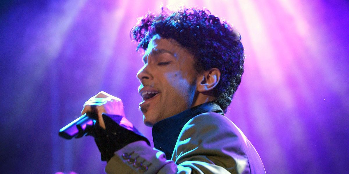 Prince holds a microphone and sings; purple lights are in the background