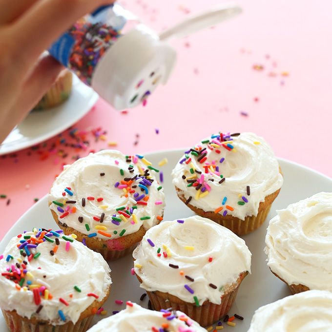 a hand adds sprinkles to cupcakes with vanilla frosting