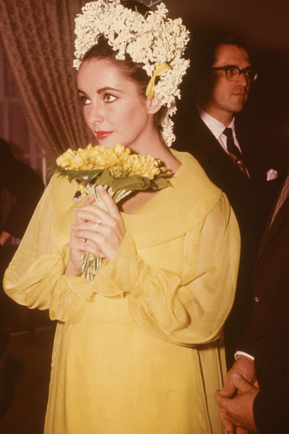 British-born actor Elizabeth Taylor, a yellow dress and floral headdress, holds a bouquet of flowers at her wedding to actor Richard Burton, March 15, 1964. (Photo by Hulton Archive/Getty Images)