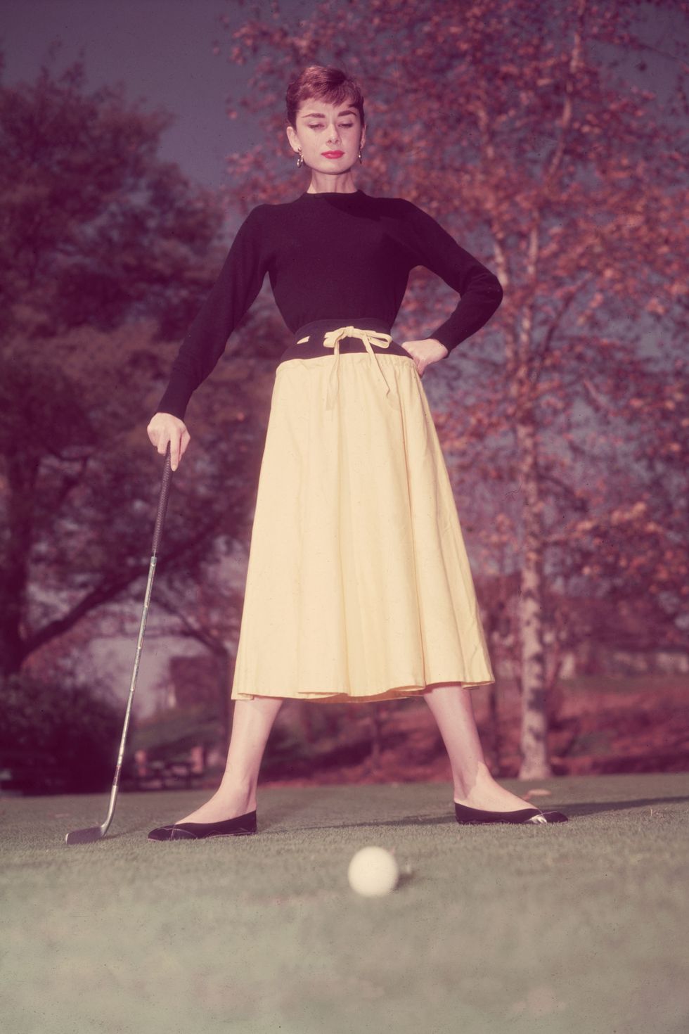 1955:  Full-length view of Belgian-born actor Audrey Hepburn (1929 - 1993) standing on a golf course, holding her putter, with a golf ball on the green in the foreground. She wears a black top and a yellow drawstring skirt.  (Photo by Hulton Archive/Getty Images)