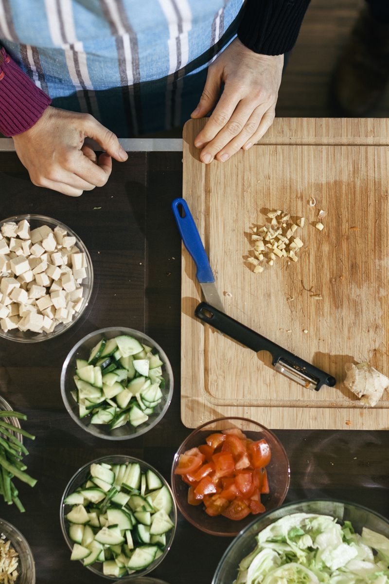 A wooden cutting board on a kitchen counter that has been used to cut vegetables during some cooking class. Around the cutting board, there are several bowls with different copped up vegetables. Hands of people using the board can also be seen.