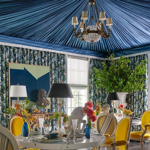 dining room with a blue tented ceiling large urns in the corner with greenery and round table with yellow chairs and lamps and a large painting over a gilded fireplace off to the left