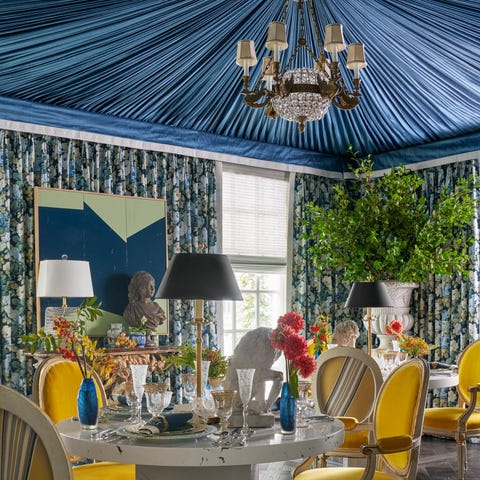 A List 2022 - ELLE DECOR dining room with a blue tented ceiling large urns in the corner with greenery and round table with yellow chairs and lamps and a large painting over a gilded fireplace off to the left