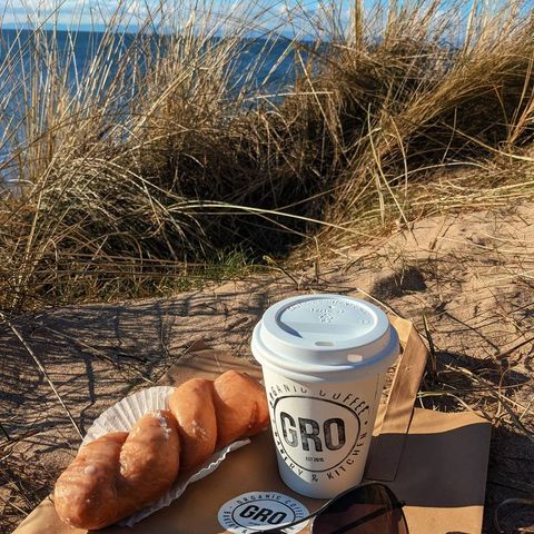a takeaway coffee cup and a pastry on sand by the sea