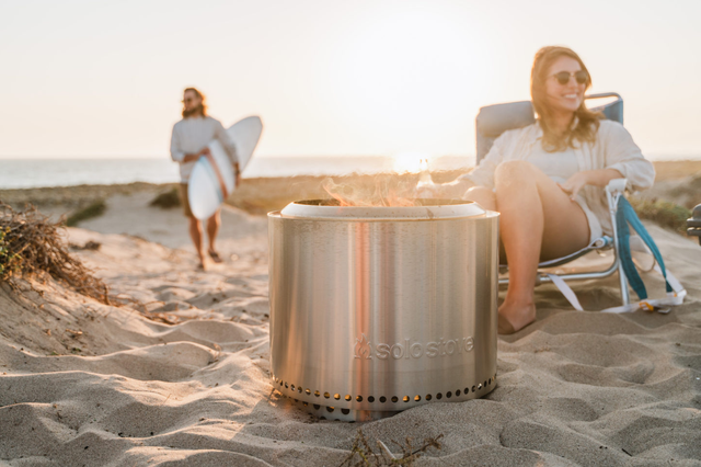solo stove portable firepit on the beach with woman in beach chair and man holding surfboard