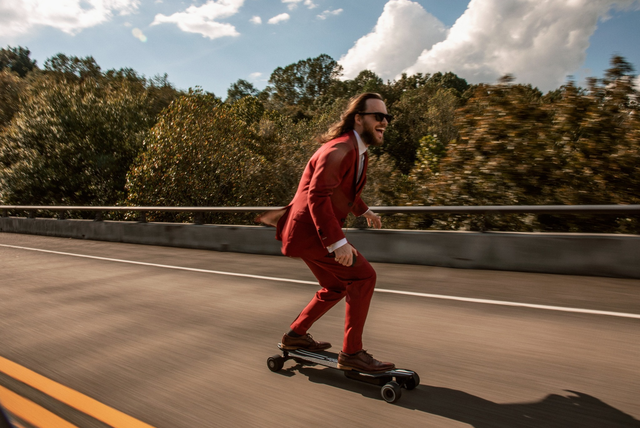 longhaired man skateboarding down street in sunglasses and red indochino suit