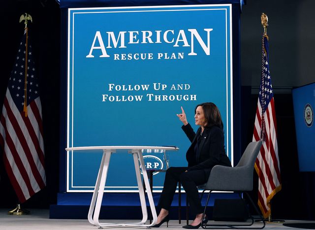 kamala harris sitting in front of a large poster for the american rescue plan
