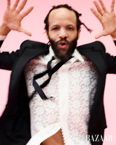 savion glover in black suit white floral lace shirt pink background