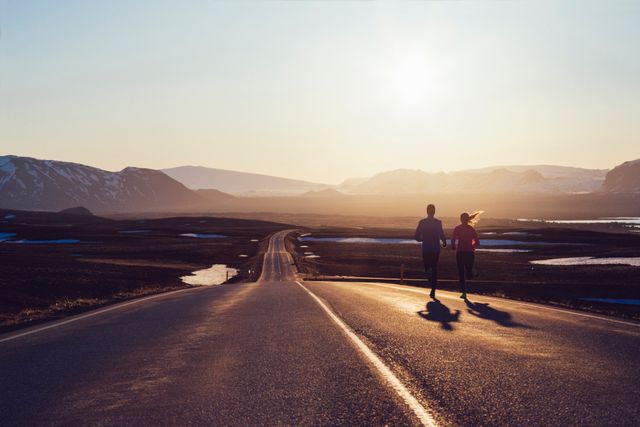 low angle view of two runners running side by side together into the distance, running along a long road heading to the mountains in iceland, beautiful early morning sunshine rising up above the distant hills