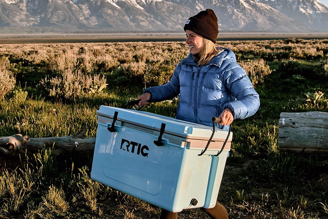 woman carrying rtic ultralight cooler across outdoor landscape with mountains in the background