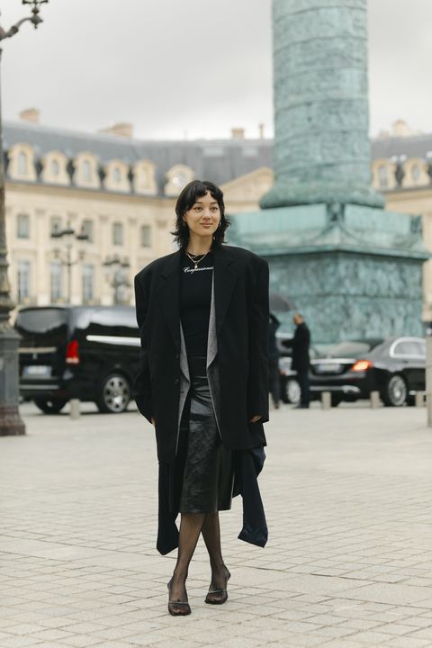 donna con capelli neri alle spalle indossa cappotto nero sandali neri gonna in pelle lunga blazer grigia e maglia nera