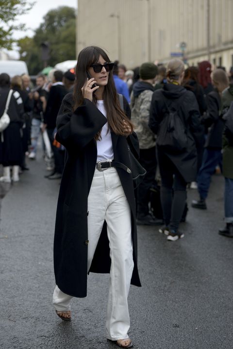 donna capelli castani che indossa occhiali da sole, cappotto nero, maglia bianca,cintura nera, pantaloni bianchi e sandali