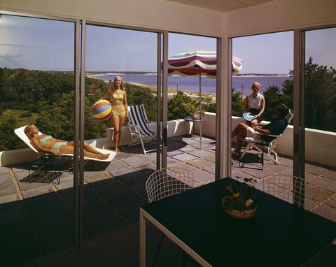 view through the windows of a corner room at the colony resort where, on the balcony, one young woman in a bikini sunbathes on a deckchair, another poses with a beach ball and a couple in casual attire talk together, wellfleet on cape cod, massachusetts, 1960s photo by aladdin color incgetty images