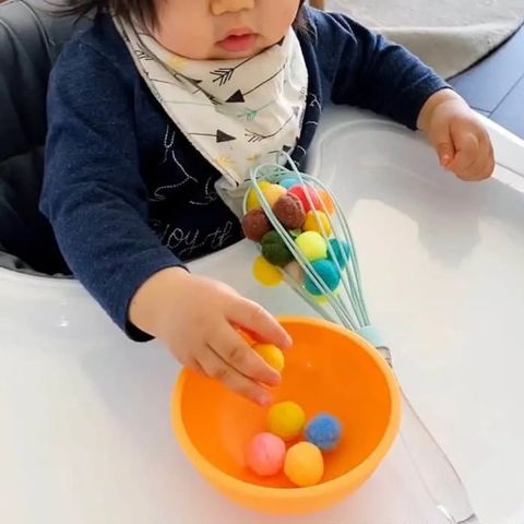 a toddler frees pom poms from inside a kitchen whisk and puts them in a bowl