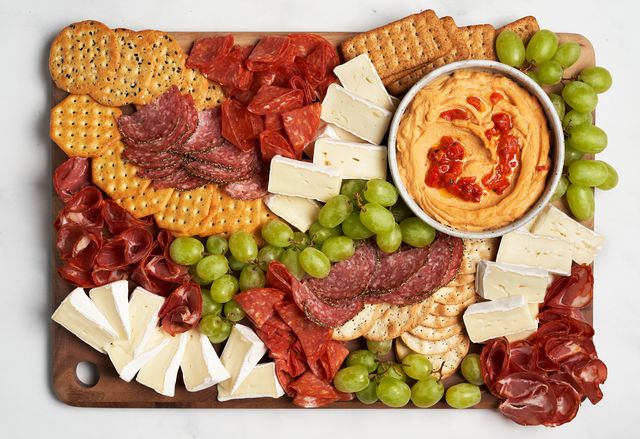 a wooden board filled with cheeses meats grapes and crackers plus a bowl of dip