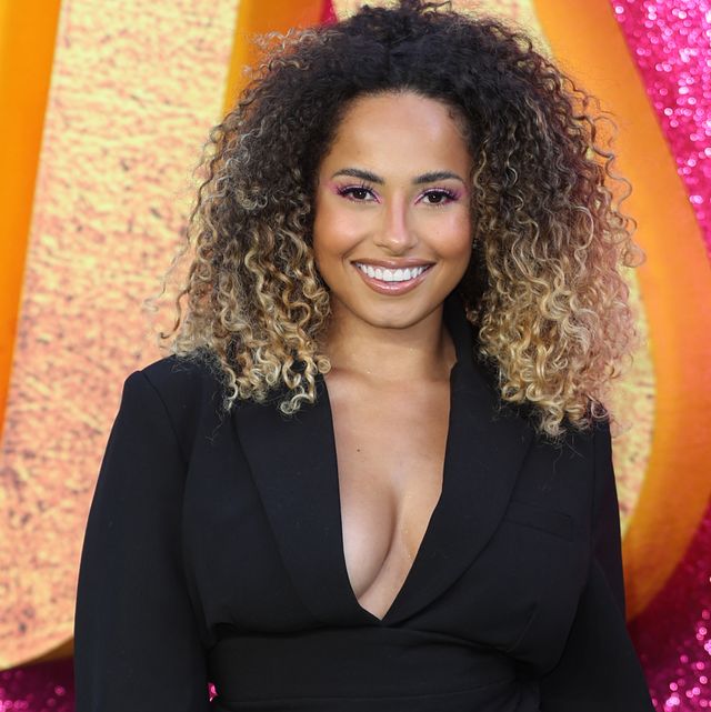amber gill standing and smiling at the camera, wearing black shorts and blazer with boots, black curly hair with blonde ends to shoulders