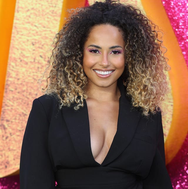 Amber Gill amber gill stands and smiles at camera, wearing black shorts and blazer with boots, shoulder length black curly hair with blonde tips