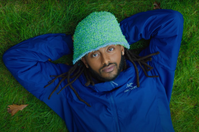 a man wearing a bucket hat and blue zip up jacket laying in the grass