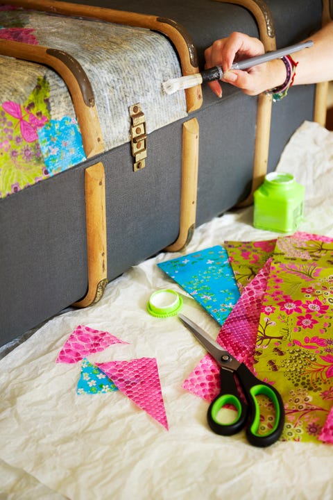 Ein Kunsthandwerker bestreicht einen Kofferraum mit Klebstoff, Neben ihr liegt ein Teil des Koffers, der bereits mit bunten Decoupage-Papieren beklebt ist's hand is painting glue over a trunk, with supplies like scissors, decorative paper and a pot of glue beside her there is a section of the trunk already affixed with brightly colored decoupage papers