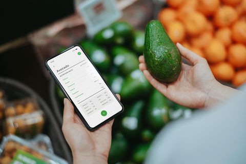 asian woman shopping for fresh organic fruits and vegetables in supermarket she is holding an avocado, using health and fitness tracker app on smartphone to check the nutrition facts and calories intake healthy eating lifestyle lifestyle and technology