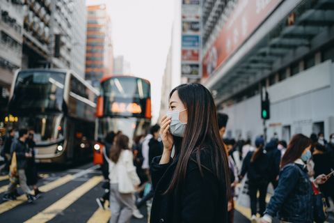 Asian woman with protective face mask crossing road in busy downtown city street against crowd of pedestrians and highrise urban buildings