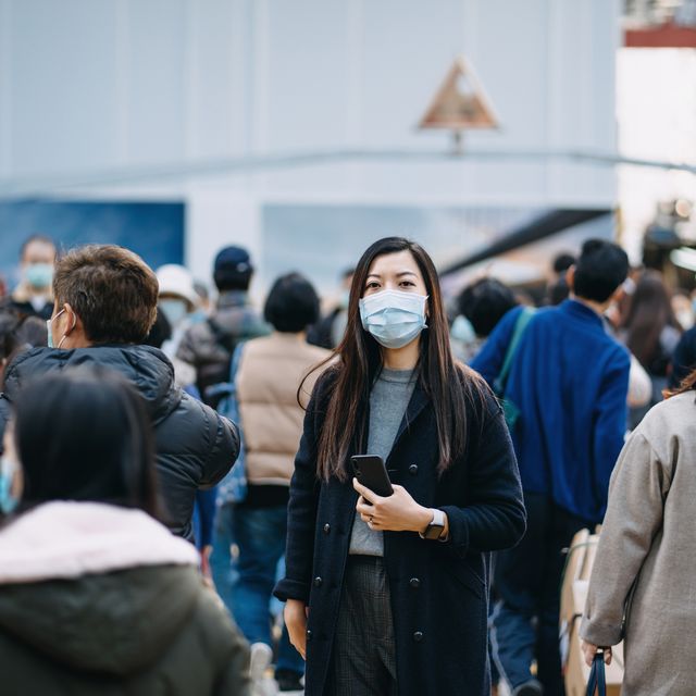 asian woman with protective face mask holding smartphone standing in the middle of busy downtown city street amidst crowd of pedestrians passing by